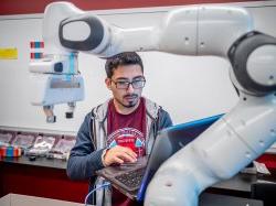 Student wearing glasses and a hoodie works on a laptop in robotics lab.