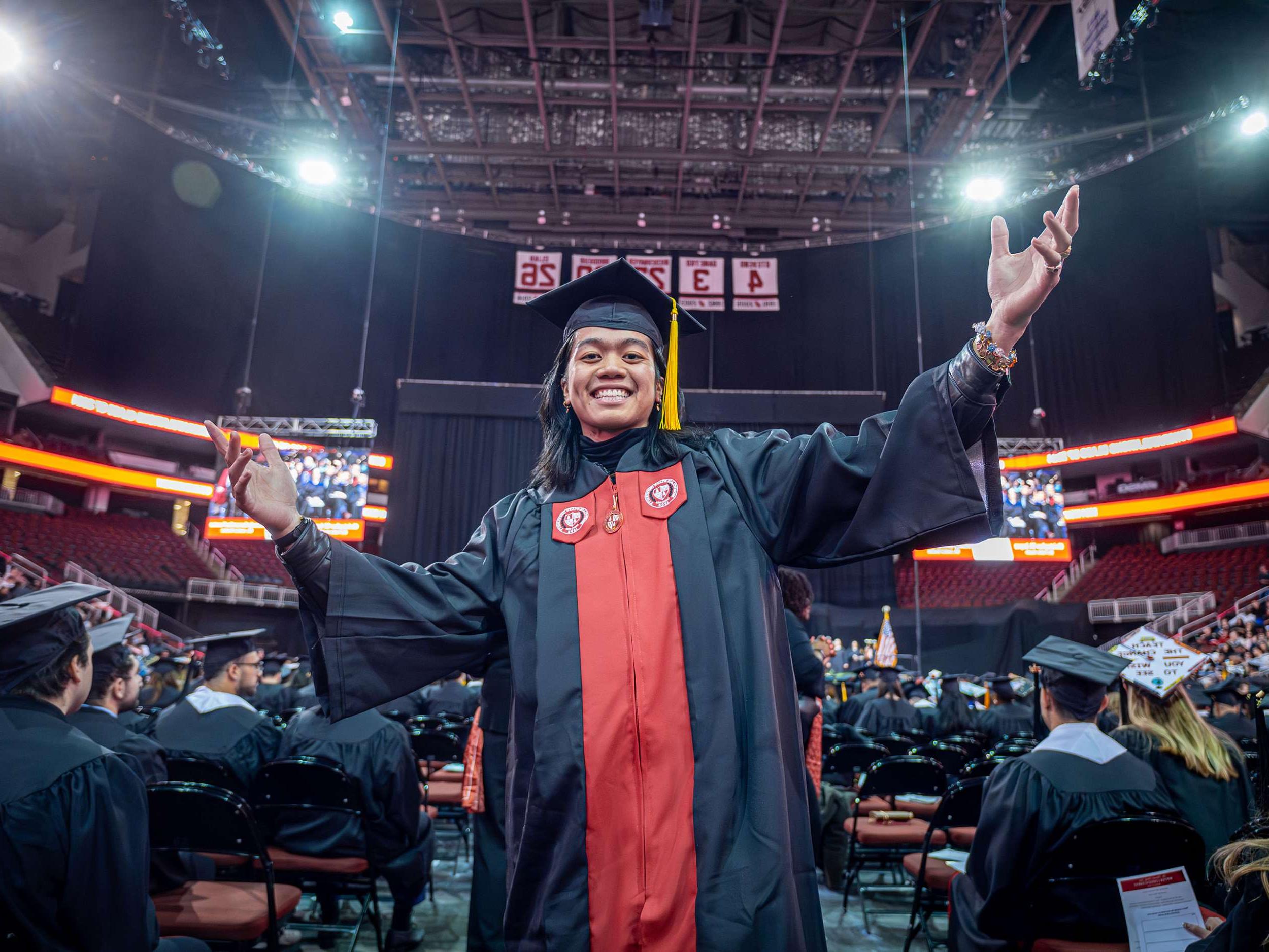 A student in a cap and gown smiles and raises his arms.