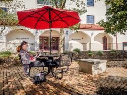 A student sitting outside of the School of Nursing and Graduate School reading a book on a sunny day under a table umbrella.