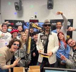 A man with a beard wearing sunglasses and tophat surrounded by a group of smiling and excited students at a college radio station.