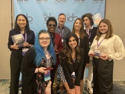 Eight people standing and smiling in front of a branded backdrop. Six of them are students who were recognized for winning national radio production awards.